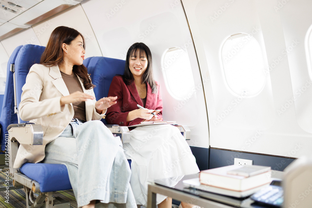 Obraz premium Two women are sitting next to each other on a plane, laughing and talking. One of them is holding a pen and a piece of paper 