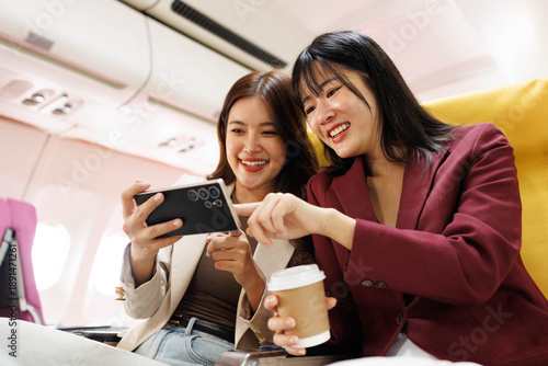 Two women sitting next to each other on an airplane, one of them looking at her phone. The other woman is writing in a notebook

