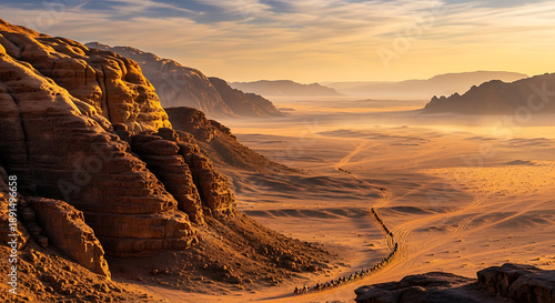 A breathtaking panoramic view of a vast desert landscape at sunset, featuring towering sandstone mountains, a winding path, and a hazy golden sky.