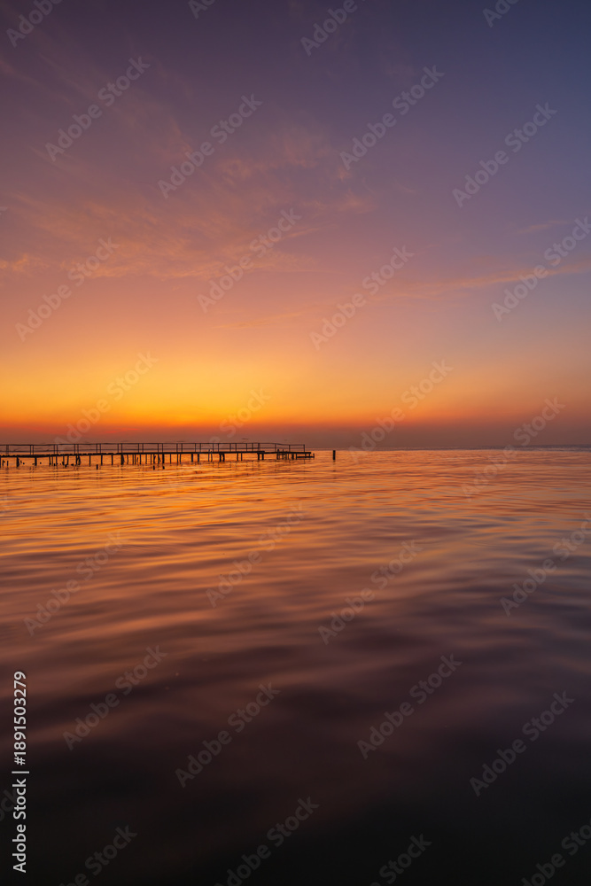 Fototapeta premium Serene Golden Sunset over Calm Sea with Wooden Pier Silhouette,A breathtaking long exposure shot of a vibrant sunset over the ocean, featuring a tranquil wooden pier stretching into the calm water. 
