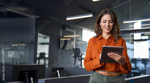 Mature adult professional business woman looking at online trade app. European businesswoman CEO holding digital tablet using fintech tab application standing at workplace in office.