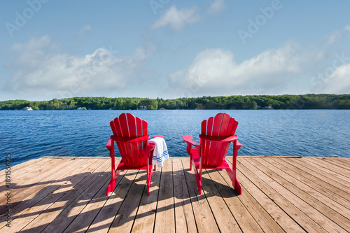 Two red Adirondack chairs on a timber dock with a blue-striped towel. Symmetrical view of an expansive blue lake and distant summer cottages.
