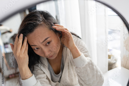 Schilderij op canvas Middle aged Asian woman looks in the mirror at her gray hair roots