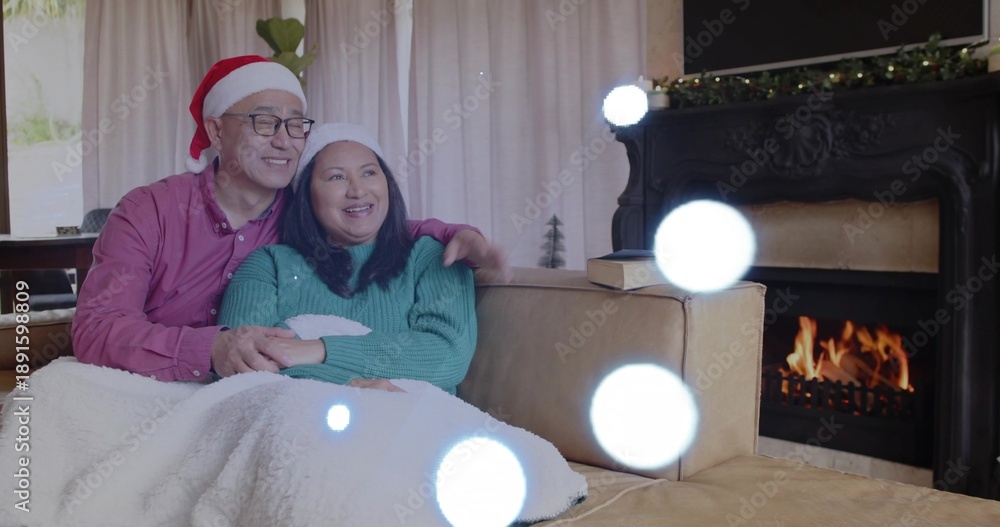 Fototapeta premium Smiling couple sitting on beige sofa at home, wearing Santa hat and teal sweater, fireplace glowing