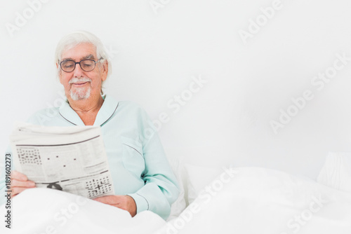 Senior man sitting in bed reading broadsheet newspaper wearing mint pajamas, glasses, copy space