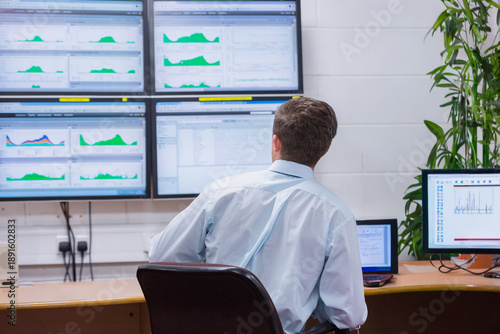 Curved wooden workstation displaying green charts on six wall screens, with potted plant near desk