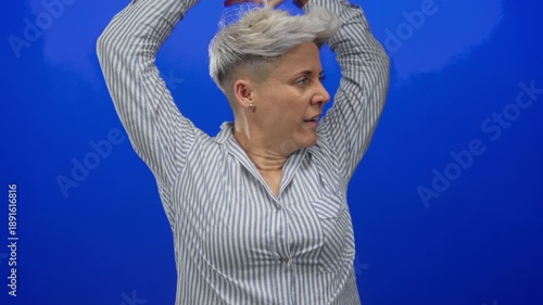 Woman in striped shirt yawning and stretching arms behind head against vivid blue studio wall; fatigue.