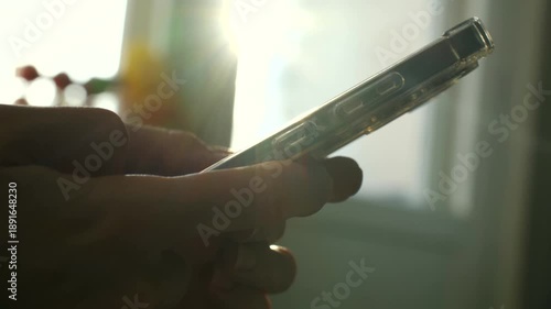 Female hands holding and using modern smartphone, scrolling, typing on touchscreen, warm morning sunlight filters through window, creating relaxed home atmosphere. Hands using smartphone.