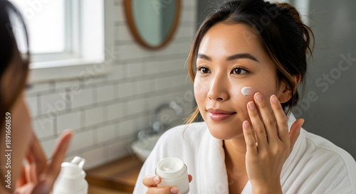 A young Asian woman applies facial cream in front of a bathroom mirror, illustrating a daily skincare routine, self-care, and healthy skin concept.