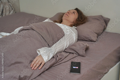 Mature caucasian woman lying awake in bed at night with phone showing 5:01 am.