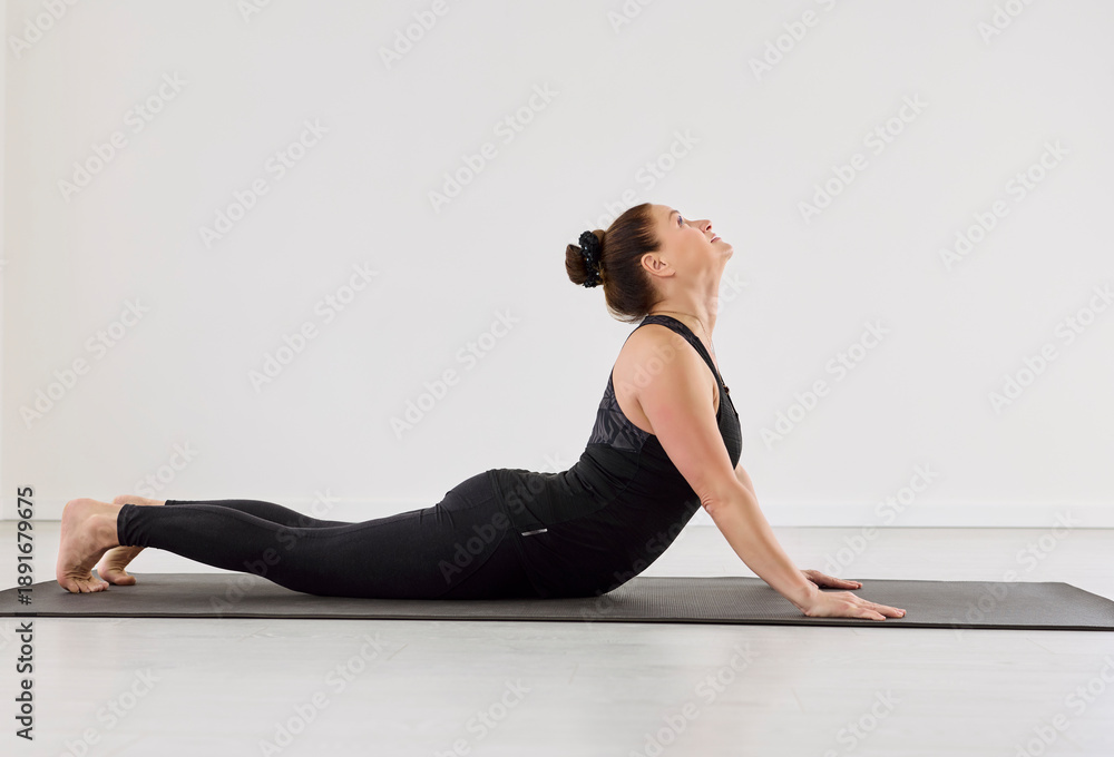 Fototapeta premium Yoga woman in cobra sphinx pose exercise. She lifts the chest on a mat in a bright studio, showing fitness form and mindful steady breathing. Concept: flexibility, wellness, focused stretch.