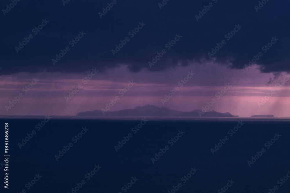 Fototapeta premium Cinematic wide shot of a distant island under a stormy purple sky. Features dramatic rain curtains and deep blue ocean. Ideal for travel, atmospheric, and background themes.