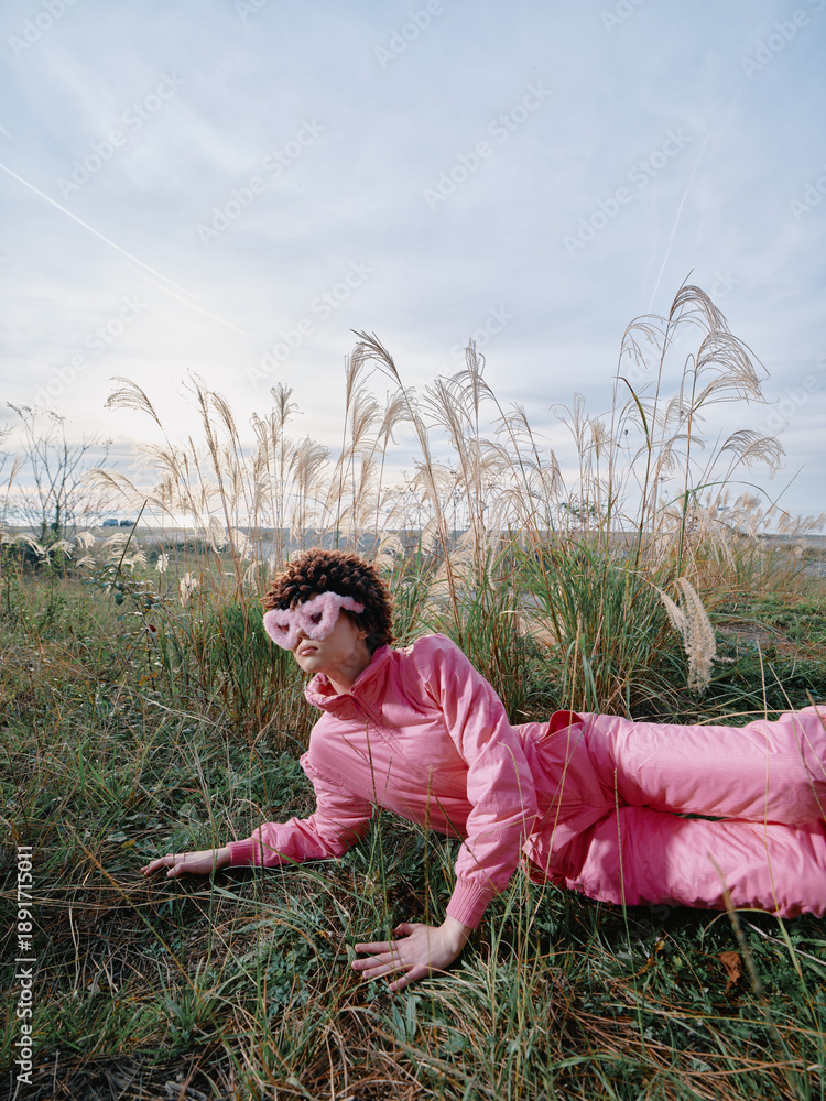 Fototapeta premium A person in a pink outfit lies on grass in an open field, relaxed pose among tall weeds, blue sky and soft light creating a casual outdoor portrait with nature