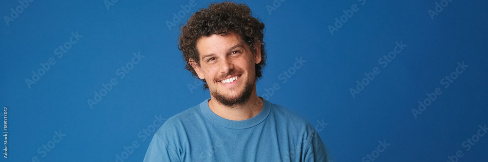 Obraz premium Smiling guy with curly hair dressed in blue t-shirt looking at the camera and waving his hand in welcoming gesture, saying hello to someone, isolated on blue background in the studio. Panoramic