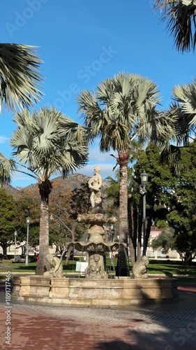 A beautiful fountain stands in a park surrounded by tall palm trees and lush greenery under a clear blue sky in Torremolinos, Malaga, Spain