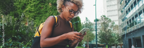 Smiling young woman in glasses, wearing brown top, uses mobile phone, chatting with friends on social networks while sitting outdoors. Panoramic