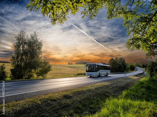 A white bus driving on the asphalt road in rural landscape at sunset