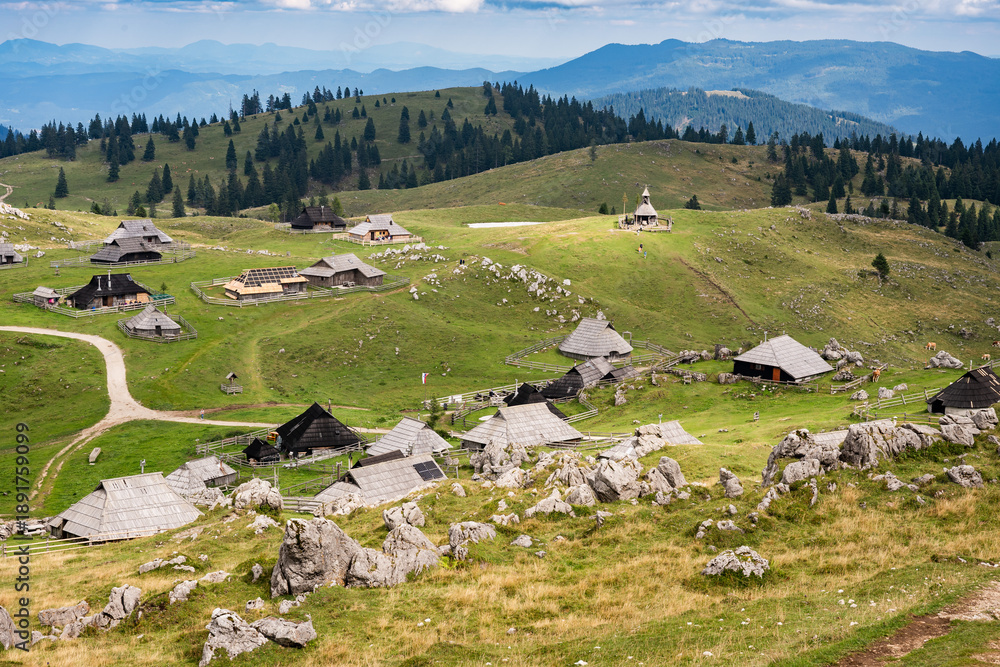 Obraz premium A wide panoramic view of a traditional shepherd village with unique wooden huts scattered across green rolling hills under a dramatic sky in Velika Planina, Slovenia.