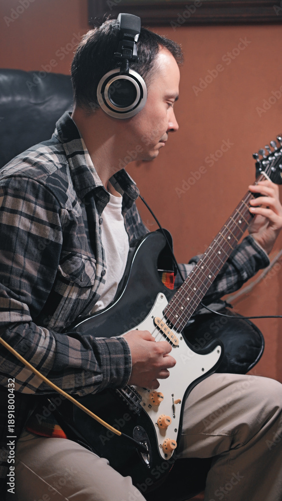Obraz premium A focused man in a plaid shirt sits while playing an electric guitar. He wears headphones, suggesting a recording session or practice. Soft, warm lighting creates a creative, introspective mood