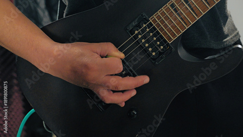 A close-up action shot of a rock musician's hand strumming a matte black electric guitar. The focused, dynamic composition captures the energy and passion of a rehearsal or live performance