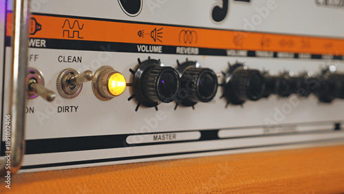 Detailed close-up of a retro guitar amplifier. The shot has a shallow depth of field focusing on the knobs and switches. A warm orange light glows, indicating the amp is on. Ideal for music themes