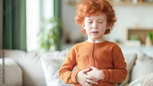Red-haired boy in orange shirt holding his stomach, standing in a bright, cozy living room, concept of stomach ache, parasites, and digestive issues in children