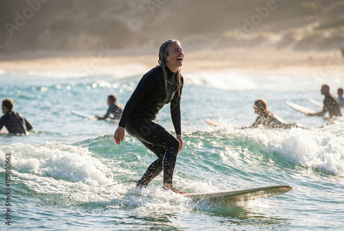 A happy surfer in a black wetsuit riding a small wave in the ocean with other surfers in the background on a sunny day.