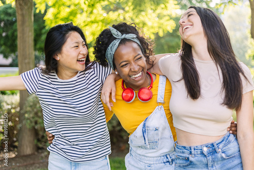 Young group of diverse teenage girls having fun together at city park, laughing while walking hugging each other. Female friendship and vacation lifestyle concept