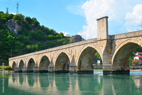 Historic stone bridge over the Drina River in Visegrad, Bosnia and Herzegovina, surrounded by green hills and calm water, famous Ottoman architecture landmark.