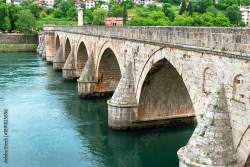 Iconic Ottoman stone bridge in Visegrad, Bosnia and Herzegovina, crossing the Drina River with arches reflected in clear green water.