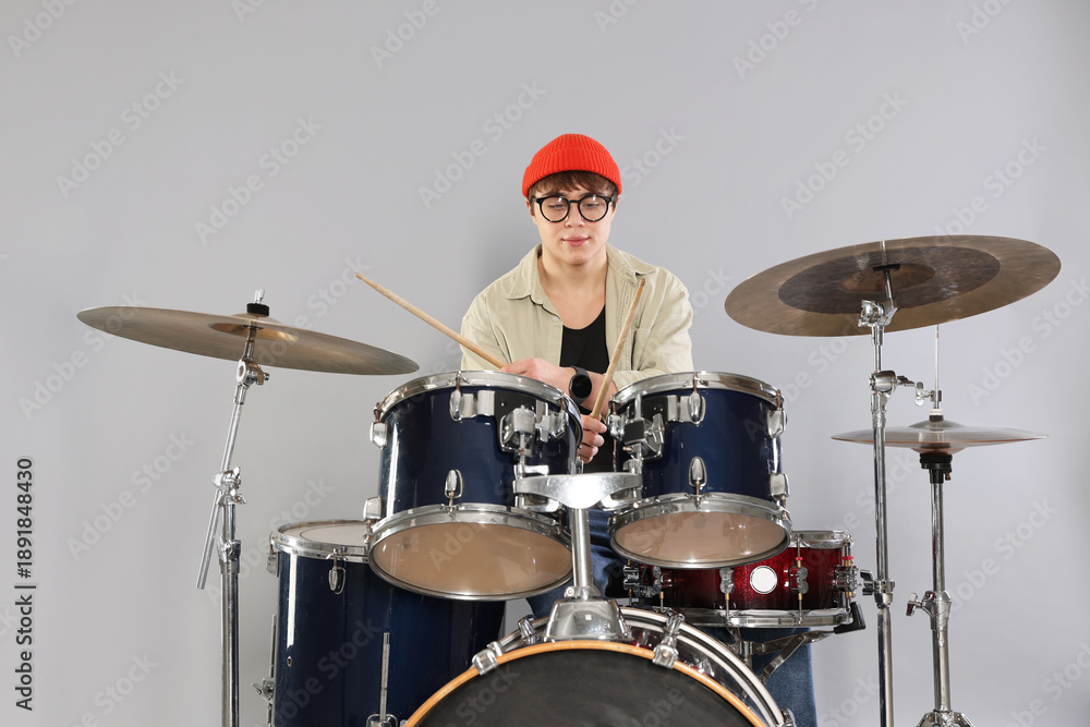 Obraz premium Teenage boy in glasses and hat with drumsticks learning how to play drums on grey background