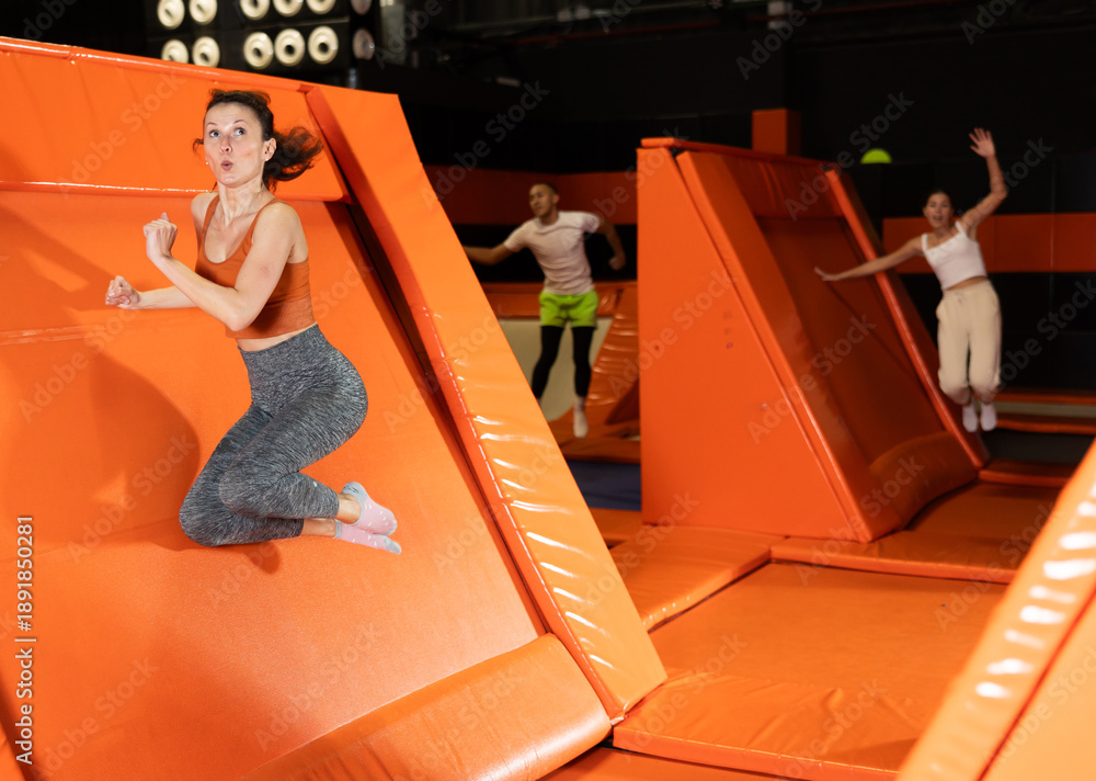 Fototapeta premium Happy emotional woman in fitness clothes jumping, pushing off from angled surface in bustling indoor trampoline park
