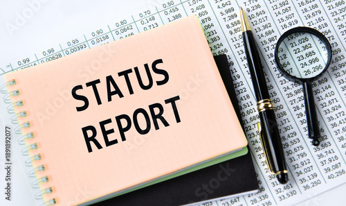 STATUS REPORT - words on a yellow notebook on a background of digital tables, a magnifying glass and a pen