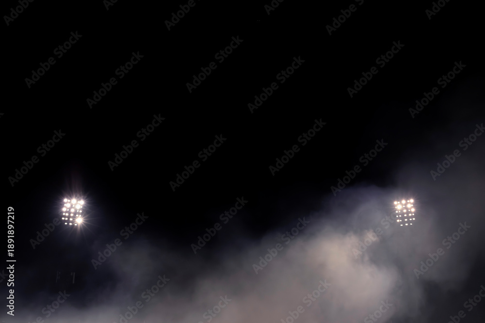 Fototapeta premium Two stadium floodlight arrays glowing through dense fog at night, symmetrical composition with smoke and mist