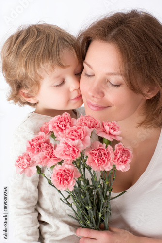 Mothers Day, Valentines Day, love holiday theme. A woman tenderly embracing a child, both wearing white clothing, against a white background. The woman holds a bouquet of pink carnations.