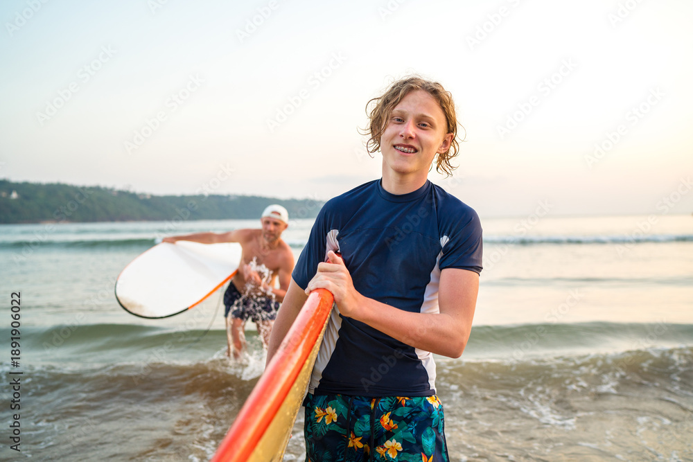 Naklejka premium Portrait of a teen boy with a surfboard after surfing with his father. They are smiling and walking out of the water. Family active vacation concept.