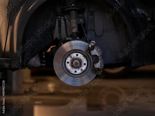Car brake disc and caliper visible with wheel removed, automotive workshop repair and maintenance scene