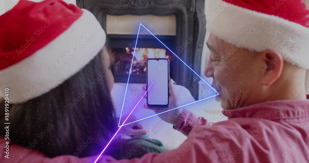 Obraz premium Sitting couple wearing Santa hats holding smartphone showing blank screen in living room, fireplace