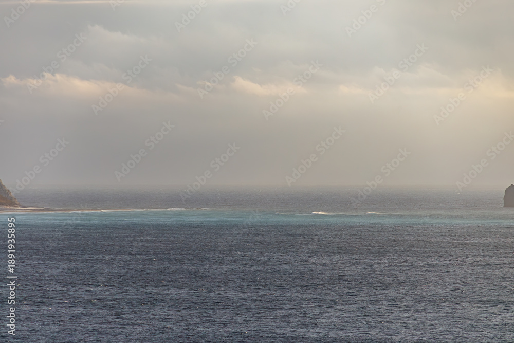 Fototapeta premium 日の出の海。 ぐんじ山展望台から見る美しい伊豆諸島の海。 沖には新島や神津島が見える。 白い石灰岩の崖、白ママ断崖の沖には無人島の早島。 乗用車を乗せた大型貨物船や漁船も見える。 式根島は東京都心から南へ160km。 外周12kmの小さな有人島。 温かい黒潮が当たり水温と水質が高い。 また、リアス式の入り組んだ入り江が多く、海水浴や釣りが盛ん。 温泉島としても有名。海際に多くの無料露天風呂がある。