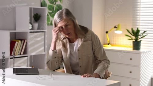 Amnesia. Senior woman with laptop trying to remember something at white table indoors