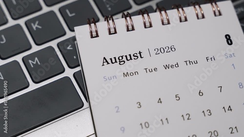 Close-up of a desk calendar showing August 2026 placed beside a laptop keyboard. Camera slowly rotates right, highlighting monthly dates and clean time management concept in a modern workspace.