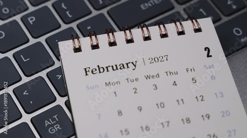 Close-up of a desk calendar displaying February 2027 on a laptop keyboard. Camera slowly moves to the right, emphasizing monthly dates and organized time management concept in a clean professional of