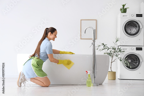 Beautiful young happy woman with bottle of detergent and rag cleaning bathtub...