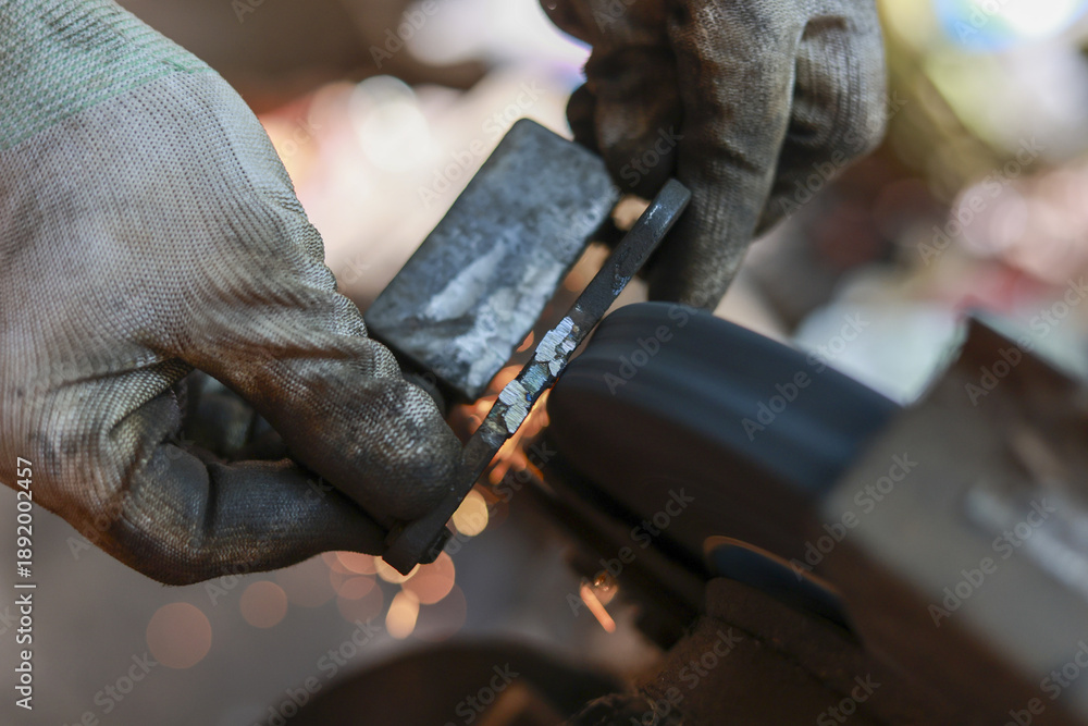 Obraz premium Intense close up of worker grinding metal with bench grinder, creating shower of hot sparks. Wearing protective gloves, craftsman carefully shapes piece of steel with focus