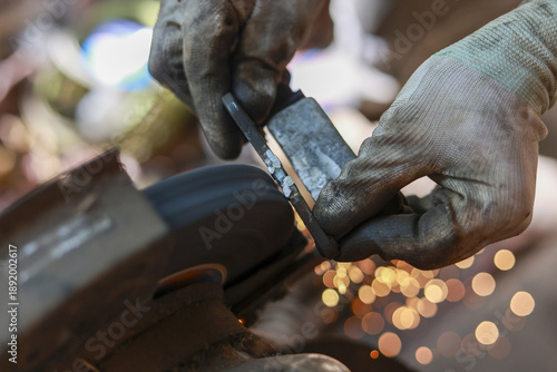 Focused blacksmith grinding metal blade with belt sander in workshop. This industrial craft shows skilled worker creating shower of sparks with careful precision