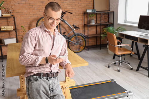 Young businessman working with mobile phone near treadmill in office