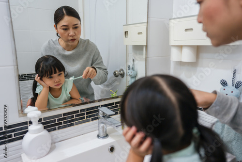 stubborn child girl refusing to brush her teeth in the bathroom. mother is trying to encourage her unhappy daughter during the morning routine, while child looks grumpy and resistant.