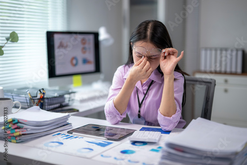 Fotografie Tired asian businesswoman rubbing her eyes at office desk after long computer wo