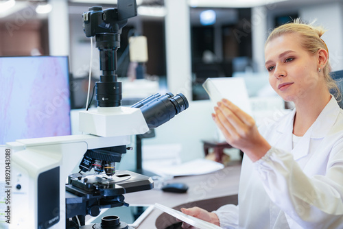 Serious Caucasian female scientist inspecting glass slide near microscope in modern lab. Young researcher checking histology sample with digital pathology monitor background.