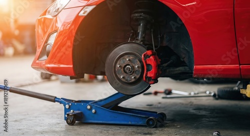 Close-up of a red car lifted by a hydraulic jack for brake system maintenance and repair in a professional automotive garage, showing detailed disc brake rotor and caliper components.
